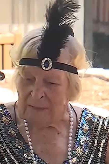 An older adult female wearing a sparkly black sequined dress with a feathered headband stands amidst blooming flowers in a garden setting during spring or summer.