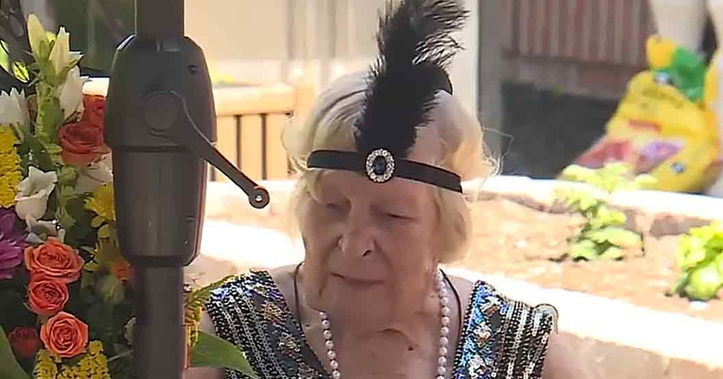 An older adult female wearing a sparkly black sequined dress with a feathered headband stands amidst blooming flowers in a garden setting during spring or summer.