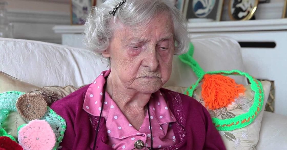 An older adult female in a cozy living room with colorful crocheted pillows on a white sofa, surrounded by home decor and framed artwork.