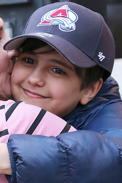 An adult female hugs a child outdoors, both wearing dark blue puffer jackets with pink and navy accents, under natural light during what appears to be autumn or spring.