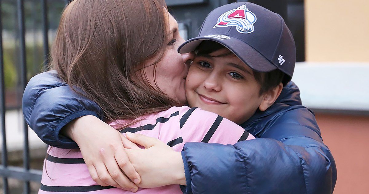 An adult female hugs a child outdoors, both wearing dark blue puffer jackets with pink and navy accents, under natural light during what appears to be autumn or spring.
