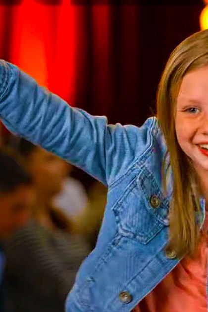 A young adult female child in a denim jacket over a pink shirt at an indoor carnival or fair with red curtains and yellow lights.