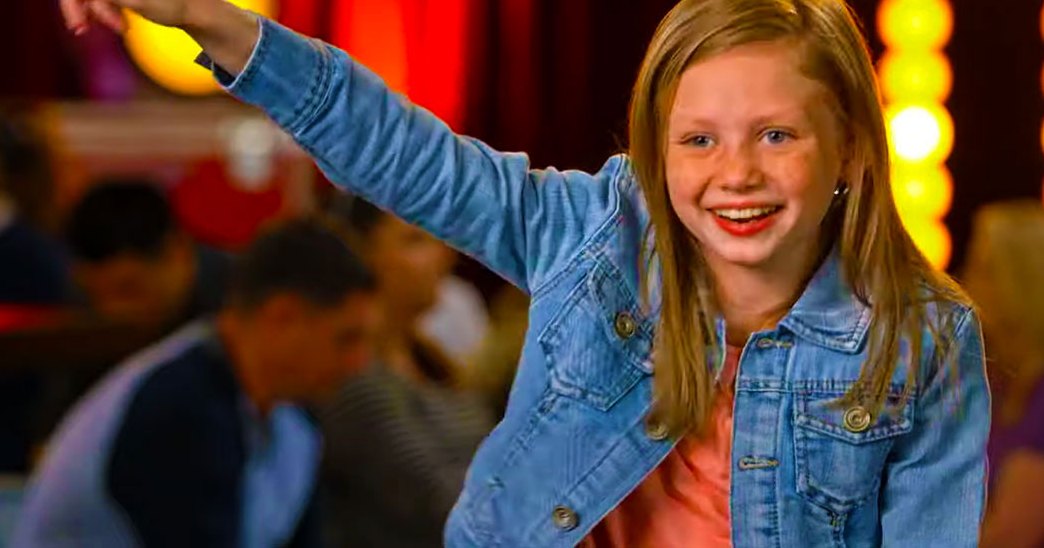 A young adult female child in a denim jacket over a pink shirt at an indoor carnival or fair with red curtains and yellow lights.