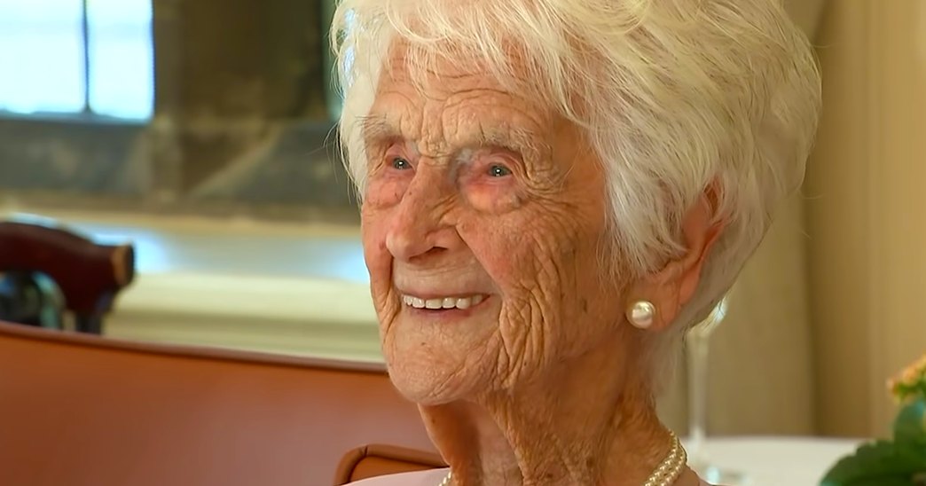 An older adult female wearing pearl earrings and a white garment with a high neckline, sitting in a neutral-colored home interior with natural light coming through a window