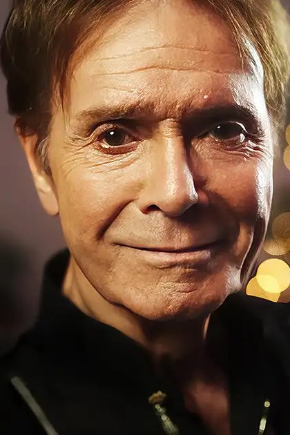 An older adult male wearing a dark, textured jacket or sweater with zippers, set against festive bokeh lights suggesting Christmas time.