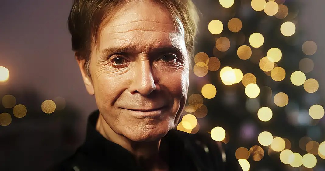 An older adult male wearing a dark, textured jacket or sweater with zippers, set against festive bokeh lights suggesting Christmas time.