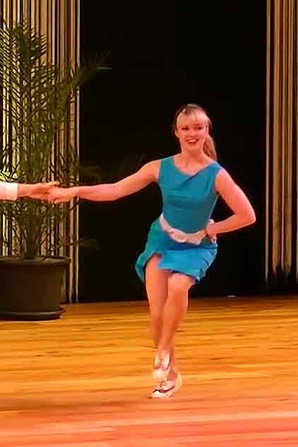A dance performance scene with young adult male and female dancers in a formal indoor setting with stage lighting, curtains, and audience presence.