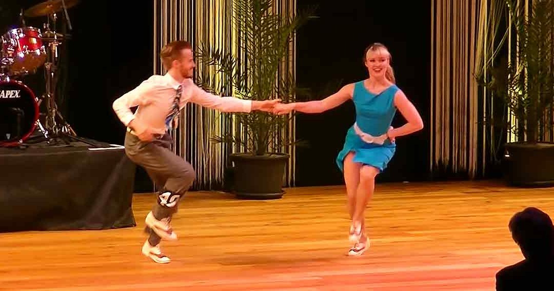 A dance performance scene with young adult male and female dancers in a formal indoor setting with stage lighting, curtains, and audience presence.