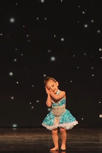 A toddler girl in a modern children's dance attire, barefoot on an indoor stage with a dark starry backdrop.