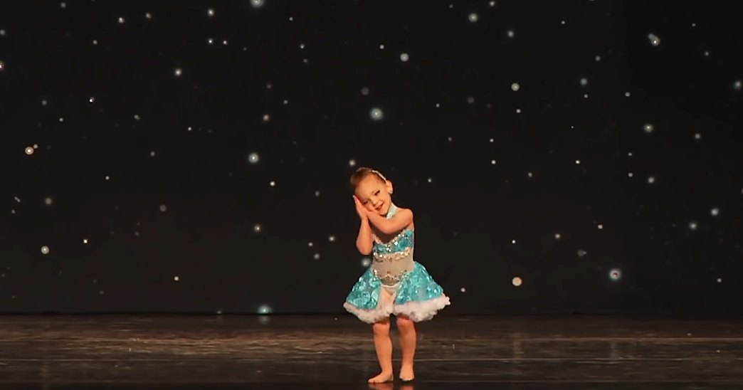 A toddler girl in a modern children's dance attire, barefoot on an indoor stage with a dark starry backdrop.