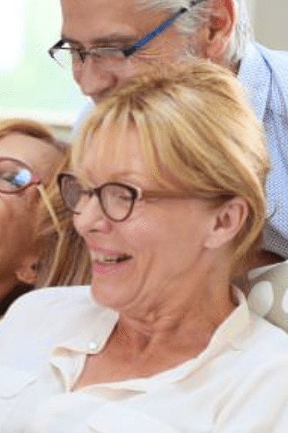Three older adults in an indoor setting, possibly a living room with patterned cushions on what seems like a sofa, interacting with each other under natural light.