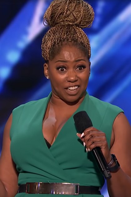 An adult female performer in a modern green sleeveless dress with a deep V-neckline, holding a microphone on an indoor stage with blue-toned geometric backdrop and stage lighting.