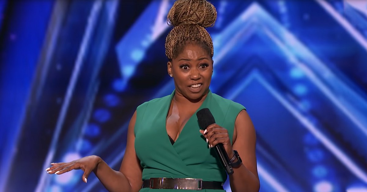 An adult female performer in a modern green sleeveless dress with a deep V-neckline, holding a microphone on an indoor stage with blue-toned geometric backdrop and stage lighting.