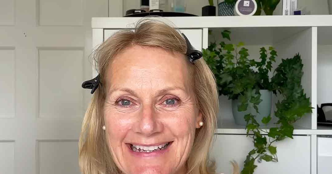 Middle-aged adult female wearing a white top with short sleeves and pearl earrings, standing in an indoor setting with green plants and containers on a white shelving unit behind her. The lighting suggests daytime.