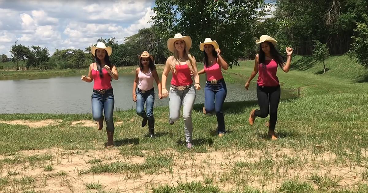 Five young adult females in a rural outdoor setting near a pond, wearing casual summer attire with pink tops and blue jeans, wide-brimmed straw hats, and various footwear under partly cloudy skies.