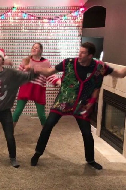 Young adults dancing in a festive Christmas living room, wearing holiday-themed outfits with red boots and patterned vests.