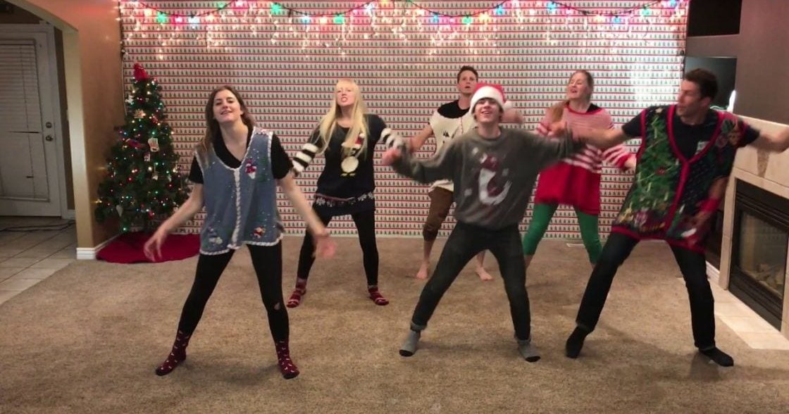 Young adults dancing in a festive Christmas living room, wearing holiday-themed outfits with red boots and patterned vests.