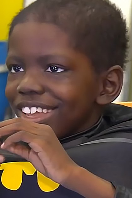 A toddler wearing a Batman costume with long sleeves and a cape, sitting indoors against yellow walls in a playful environment.