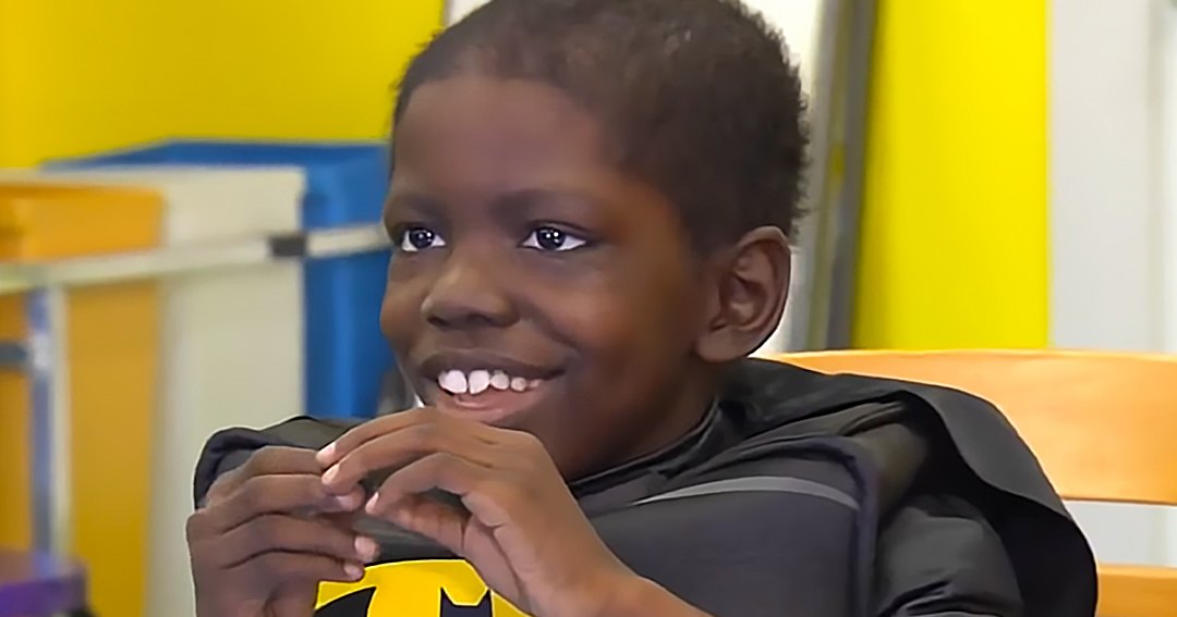 A toddler wearing a Batman costume with long sleeves and a cape, sitting indoors against yellow walls in a playful environment.
