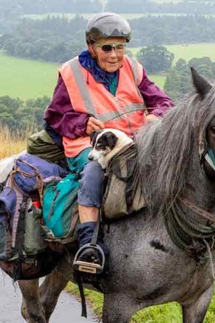 An older adult female riding a draft horse in a rural landscape with late summer or early autumn foliage and midday light.
