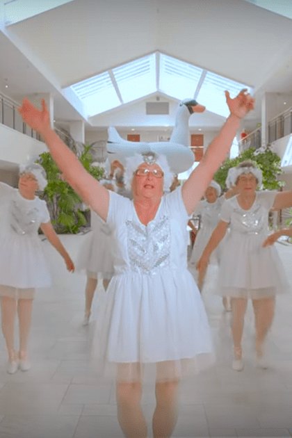 An older adult male with gray hair and glasses wearing a dark turtleneck shirt indoors; background includes white walls, skylight, plants, and a group of women in white tutus and ballet shoes suggesting a dance class or performance.