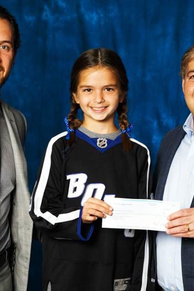 A young girl in a hockey jersey holds a check, flanked by two adults dressed formally against a plain blue backdrop.
