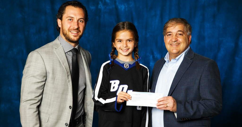 A young girl in a hockey jersey holds a check, flanked by two adults dressed formally against a plain blue backdrop.