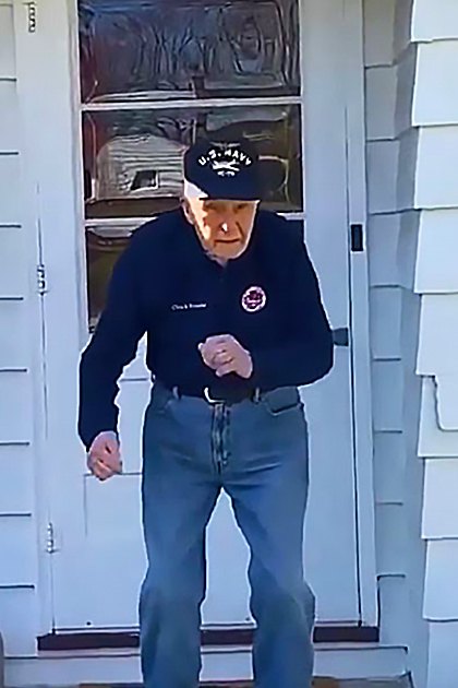 A middle-aged man wearing a U.S. Navy zip-up sweatshirt and jeans, standing outside near a house with white siding in clear daylight.
