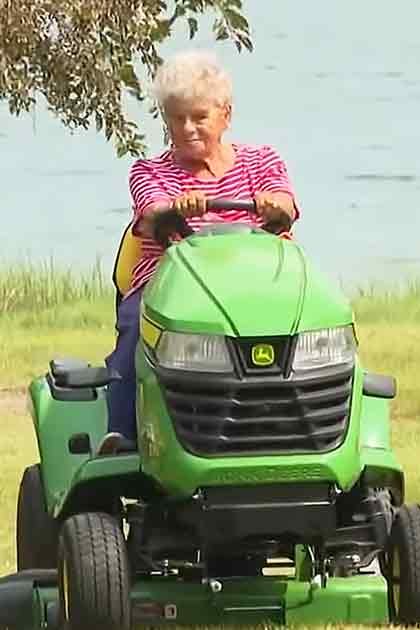 An older adult woman riding a lawn mower near water with greenery and trees in the background, wearing a red and white striped shirt.