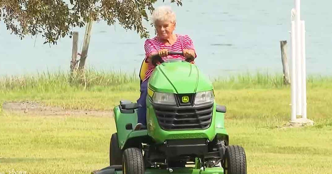 An older adult woman riding a lawn mower near water with greenery and trees in the background, wearing a red and white striped shirt.