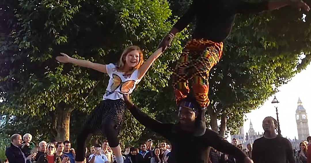 A street performer with a young adult female child in London, England, surrounded by onlookers and iconic Big Ben clock tower in the background during late afternoon or early evening.