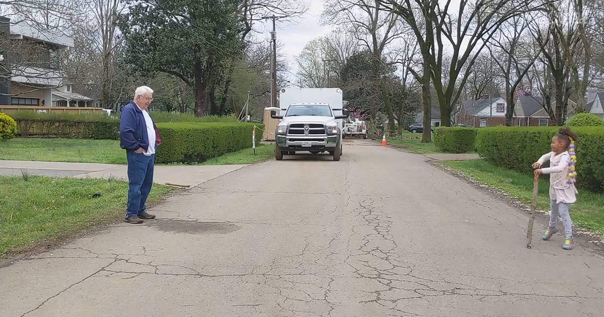 An older adult male in a blue corduroy jacket stands on a suburban residential driveway with a white pickup truck and trailer, surrounded by greenery under soft late afternoon light.