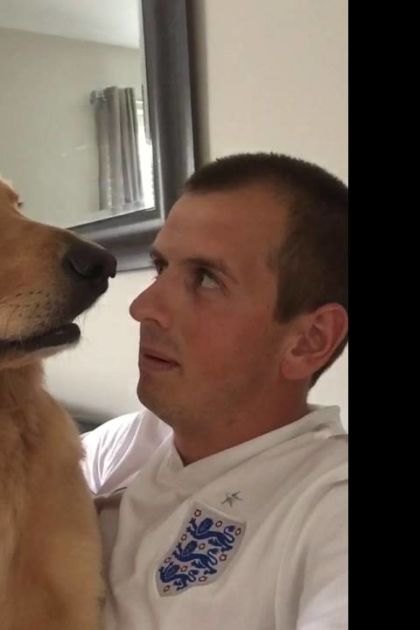 Adult male with Golden Retriever in indoor setting, white t-shirt with blue and red logo, light golden-brown dog fur, natural daylight.