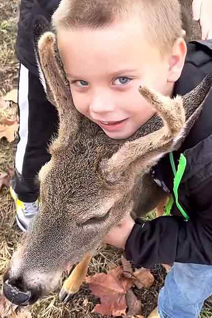 A young child and a white-tailed deer in an autumnal outdoor setting with fallen leaves and green grass.