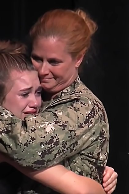 Two individuals embrace in front of a decorated Christmas tree, with one wearing a dark dress or uniform and the other in a camouflage military-style shirt. The setting appears to be indoors during the holiday season.