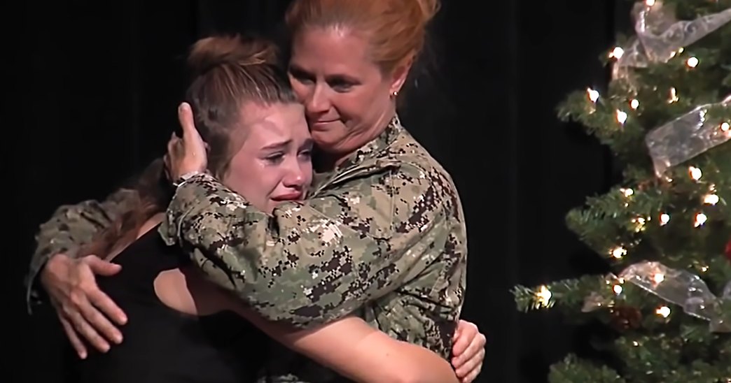 Two individuals embrace in front of a decorated Christmas tree, with one wearing a dark dress or uniform and the other in a camouflage military-style shirt. The setting appears to be indoors during the holiday season.
