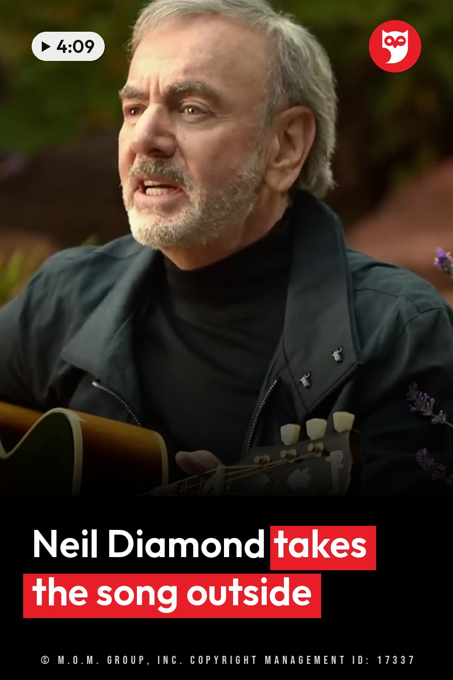 Neil Diamond, gray-haired and bearded, sings and plays an acoustic guitar outdoors near purple flowers. A headline runs in white letters across the lower third.