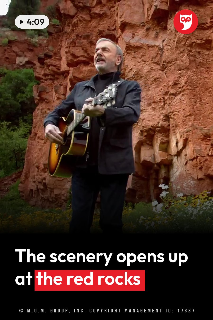 Neil Diamond plays an acoustic guitar while standing in front of a red rock cliff, with wildflowers nearby. A headline runs in white letters across the lower third.