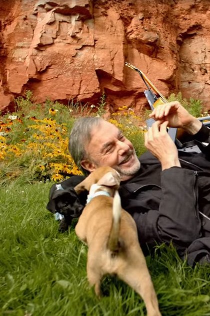 A middle-aged adult male wearing a black jacket outdoors near red rock formations and wildflowers, interacting with two light brown puppies.