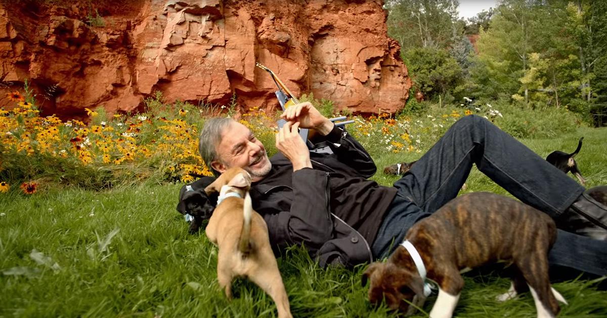 A middle-aged adult male wearing a black jacket outdoors near red rock formations and wildflowers, interacting with two light brown puppies.