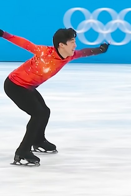 A young adult male figure skater in a red long-sleeve skating costume competes on an indoor ice rink during the Olympic event under bright, even lighting.