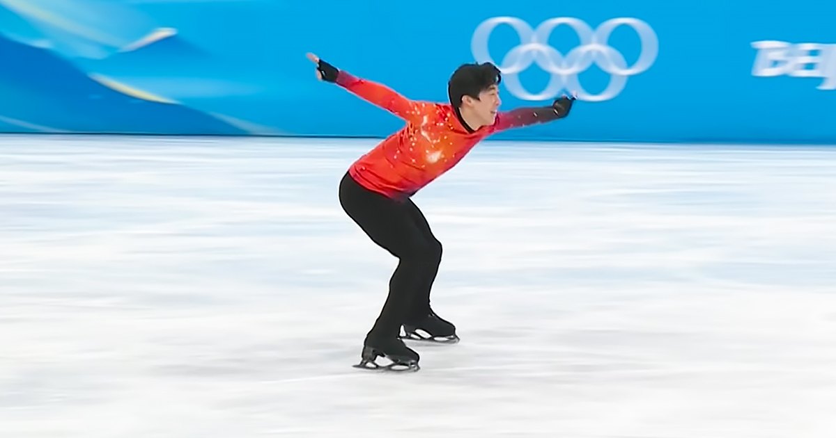A young adult male figure skater in a red long-sleeve skating costume competes on an indoor ice rink during the Olympic event under bright, even lighting.