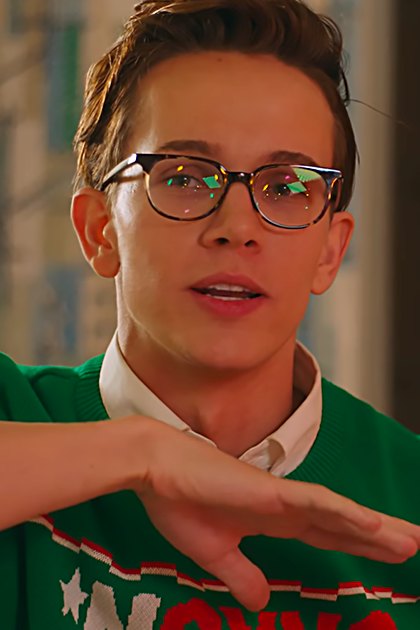 A young adult male in a relaxed, slightly oversized green sweater with white and red accents stands indoors under artificial evening lighting.