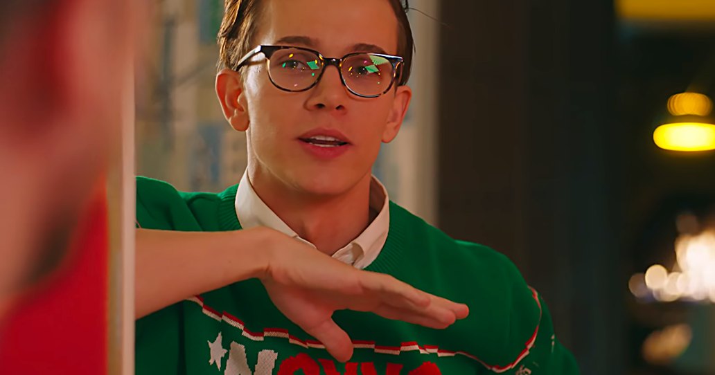 A young adult male in a relaxed, slightly oversized green sweater with white and red accents stands indoors under artificial evening lighting.
