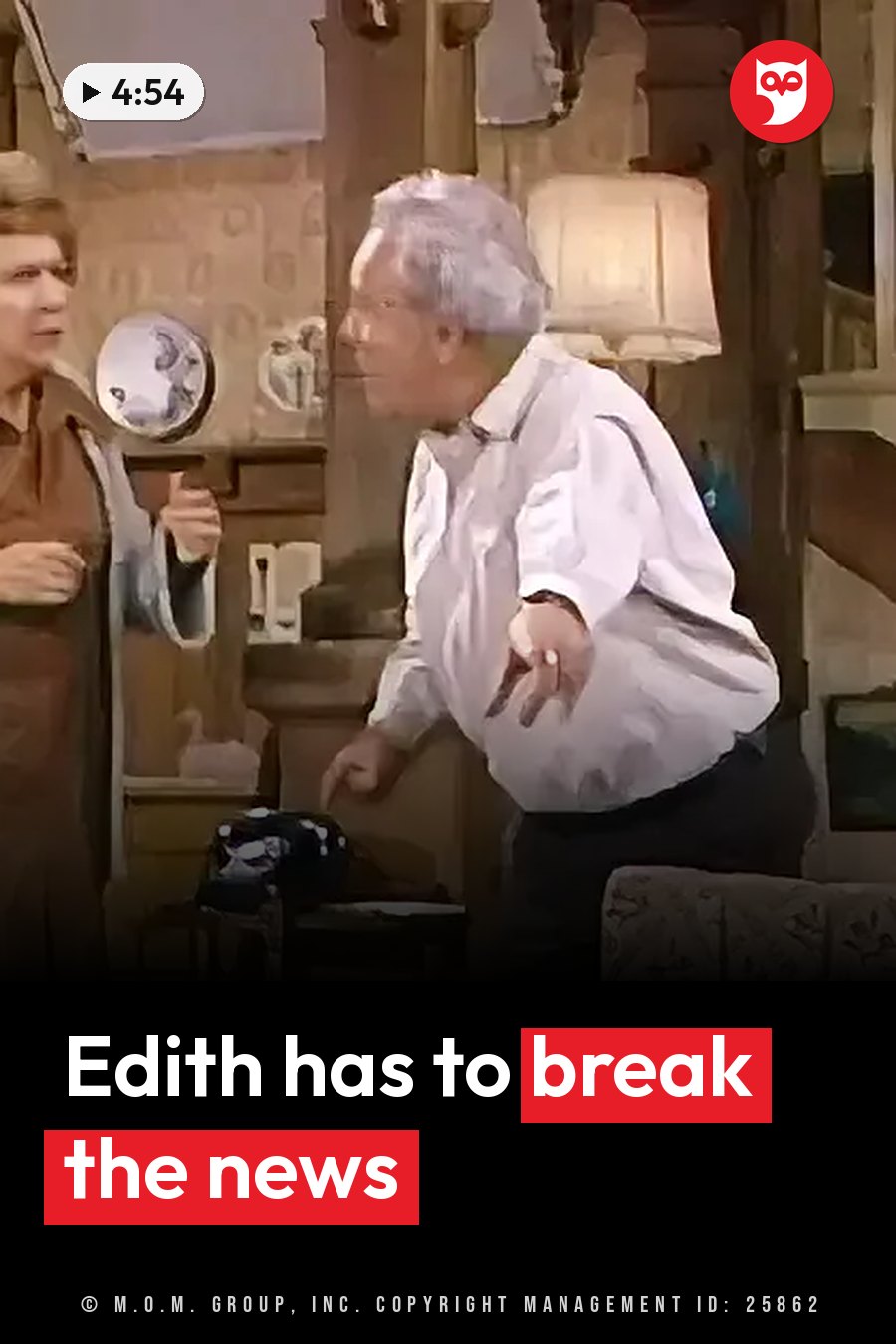 Two people stand indoors near a lamp and a decorative plate, one gesturing while speaking to the other. A headline runs in white letters across the lower third.
