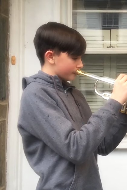 A child plays the trumpet outside a house with older adults watching from inside; scene takes place in spring or summer with white-painted shutters and greenery visible.