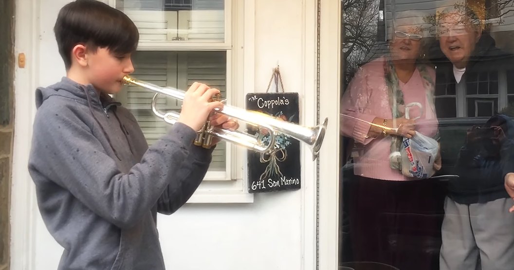 A child plays the trumpet outside a house with older adults watching from inside; scene takes place in spring or summer with white-painted shutters and greenery visible.