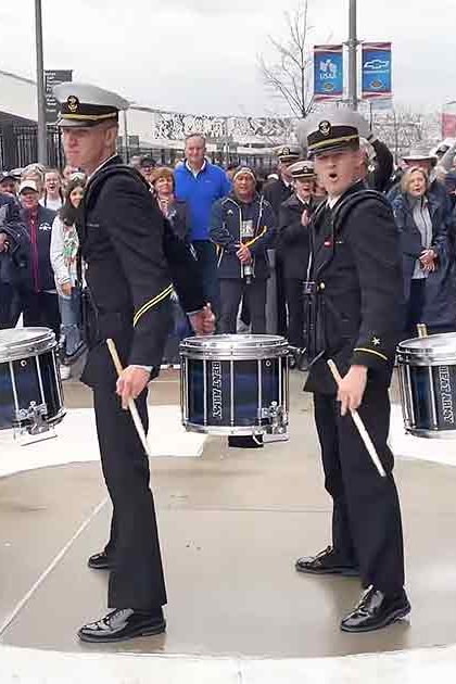 A marching band at an outdoor event near a stadium with young adult to middle-aged male and female members in military-style uniforms holding drumsticks.