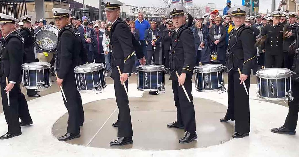A marching band at an outdoor event near a stadium with young adult to middle-aged male and female members in military-style uniforms holding drumsticks.