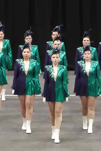 Young adult female dancers performing in a studio setting, wearing shiny green jackets with white accents and matching skirts, white ankle boots, and feathered headpieces.